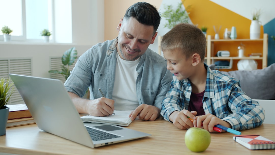 Young adult man is drawing in notebook having fun with happy child at home sitting at table with laptop together. Indoor activities and family concept.