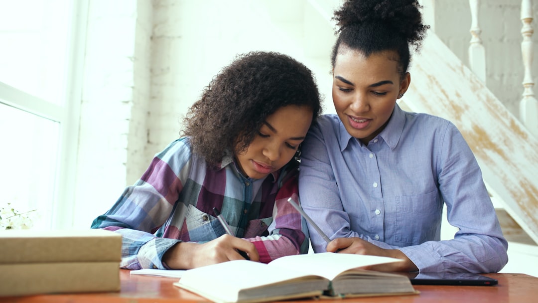 Teenage curly haired mixed race young girl sitting at the table concentrating focused learning lessons and her elder sister helps her studying at home